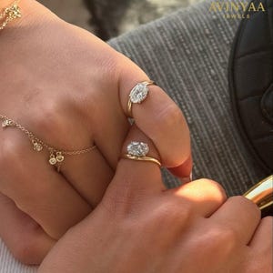 Peut inclure: Deux bagues en or avec des pierres précieuses de forme ovale sont présentées sur des doigts. Un bracelet en or délicat avec de petits charms est également visible. Les bagues sont serties dans des montures en or, mettant en valeur les pierres scintillantes.