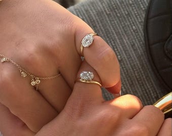 Può includere: Due anelli d'oro con pietre preziose taglio ovale sono mostrati su dita. È visibile anche un delicato braccialetto d'oro con piccoli ciondoli. Gli anelli sono incastonati in montature d'oro, mettendo in risalto le pietre scintillanti.