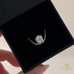 Puede incluir: Un anillo de plata con un diamante de talla redonda, presentado en una caja de joyería negra. El diamante brilla, reflejando la luz. La caja está abierta, mostrando el anillo sobre un fondo oscuro, resaltando su brillo.