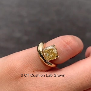 Pode incluir: Um anel de ouro com um grande diamante amarelo de corte almofada cultivado em laboratório. A banda do anel curva-se ao redor do dedo. O texto "3 CT Cushion Lab Grown" é visível.