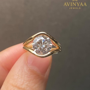 Op de afbeelding: Een gouden ring met een grote, ronde, heldere edelsteen. De ring heeft een uniek, open ontwerp dat de steen vasthoudt. Het logo "AVINYAA JEWELS" is rechtsboven zichtbaar.