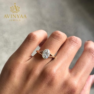 Op de afbeelding: Een gouden ring met een ovale, heldere edelsteen. De ring heeft een split-schacht ontwerp. De merknaam "AVINYAA JEWELS" staat in de linkerbovenhoek. De ring zit aan een hand.