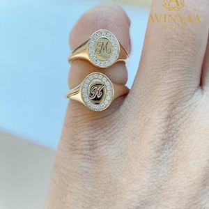 Könnte beinhalten: Zwei goldfarbene Siegelringe, jeweils mit einem diamantbesetzten ovalen Gesicht. Ein Ring zeigt den Buchstaben "M", der andere "K". Die Ringe werden an einem Finger präsentiert. Die Ringe sind von der Marke "AVINYAA JEWELS".