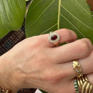 Op de afbeelding: Een gouden ring met een ovale, donkerrode edelsteen in het midden, omringd door een halo van kleinere, heldere edelstenen. De ring wordt gedragen aan een hand met andere gouden ringen en een armband. De merknaam "AVINYA" is zichtbaar.