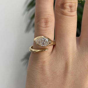 Puede incluir: Un anillo bypass de oro con un diamante en forma de pera en un engaste de bisel. La banda es lisa y pulida, envolviendo el dedo con un diseño abierto. El texto AVINYAA JEWELS es visible en la esquina superior derecha de la imagen.