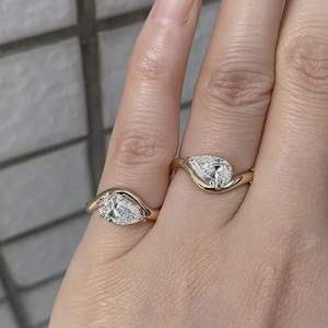 May include: Two gold rings, each featuring a single pear-shaped clear gemstone set in a smooth, curved band. The rings are worn on adjacent fingers, showcasing the polished yellow gold metal and the faceted, brilliant-cut stones.
