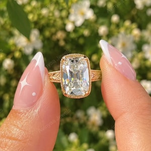 May include: A gold ring featuring a large, rectangular cushion-cut clear gemstone set in a milgrain-edged bezel. The band displays intricate engraved leaf-like patterns along the sides, providing a detailed texture to the metal setting.