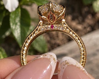 Op de afbeelding: Een gouden ring met een grote, ronde, gefacetteerde middensteen in een decoratieve, bloembladachtige zetting. De band heeft een textuur met een touwachtig patroon aan de zijkanten, geaccentueerd met kleine, heldere steentjes en een rode steen.