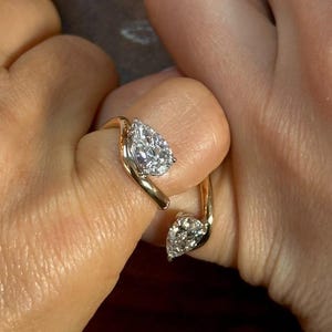 Puede incluir: Un anillo de oro con dos diamantes en forma de pera. El diseño del anillo se curva alrededor del dedo, con un diamante en la parte superior y otro en la inferior. El fondo es neutro.