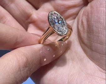 May include: A gold ring featuring a large, oval-cut diamond. The diamond is set in a classic four-prong setting, and the band is a simple, polished gold. The ring is held up by a hand, showcasing the ring's sparkle and design.