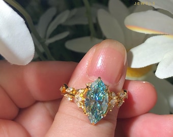 May include: A gold ring featuring a marquise-cut, light blue gemstone. The ring has a floral design with small, clear accent stones. The brand name "AVINYAA JEWELS" is visible in the background.