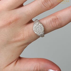 May include: A diamond ring with a leaf-shaped design, set with numerous small diamonds. The ring is on a finger. The background is a plain, neutral colour. The ring is from Avinyaa Jewelry.