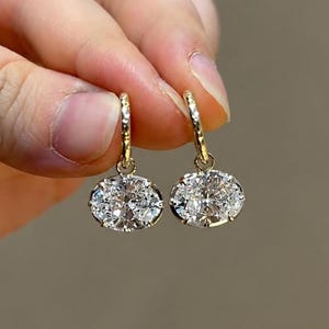May include: A pair of gold hoop earrings featuring oval-cut clear gemstones suspended from textured gold hoops. Each gemstone is secured in a prong setting, reflecting light with multiple facets. The earrings are crafted from polished yellow gold.