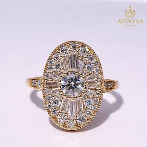 Puede incluir: Un anillo de oro con una gran pieza central ovalada. El centro presenta una piedra preciosa redonda y transparente rodeada de piedras más pequeñas, transparentes, de talla baguette y redondas. La banda y el engaste del anillo son de oro. El texto "AVINYAA JEWELS" está en la esquina superior derecha.