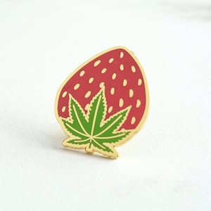 May include: A gold-toned metal enamel pin shaped like a strawberry. The fruit is red with small yellow seed-like dots, and the top features a green leaf shaped like a cannabis leaf with defined veins and pointed edges, outlined in gold metal.