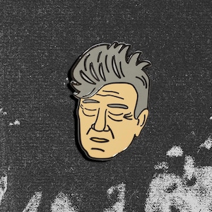 Puede incluir: Un pin de esmalte metálico con una ilustración estilizada de una persona con cabello gris grueso peinado hacia atrás y tez beige claro. El rostro tiene ojos cerrados, cejas definidas, una nariz prominente y líneas sutiles en la boca y frente.