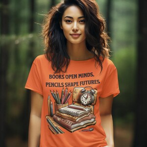 May include: Orange t-shirt with the text "BOOKS OPEN MINDS, PENCILS SHAPE FUTURES." The graphic includes books, pencils, and an alarm clock. The person is wearing gray pants and is standing in front of a blurred green background.