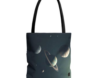 Op de afbeelding: Een zwarte tote bag met een ruimte-thema print van een geringde planeet, een grote gestreepte planeet en verschillende kleine manen tegen een donkerblauwe en zwarte sterrenhemel. De tas heeft twee stevige zwarte schouderbanden.