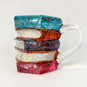 以下が含まれることがあります： カラフルなガラス製のマグカップで、本を積み重ねた形をしています。マグカップは透明な持ち手があり、青、オレンジ、紫、白、赤の色合いの本で構成されています。一番上の本には「LOVE」の文字が刻まれています。