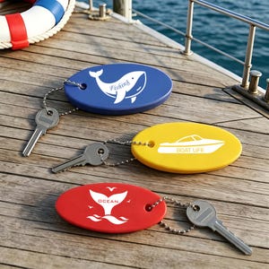 May include: Three oval floating keychains with metal ball chains and keys. The blue one features a white whale graphic and the text Fishing. The yellow one shows a white boat graphic and the text BOAT LIFE. The red one displays a white whale tail graphic and the text OCEAN.