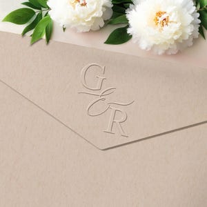 Könnte beinhalten: Ein beigefarbener Umschlag mit den geprägten Initialen "G & R" wird gezeigt. Zwei weiße Pfingstrosen mit gelben Zentren und grünen Blättern befinden sich im Hintergrund. Der Umschlag ist angewinkelt und zeigt seine strukturierte Oberfläche.