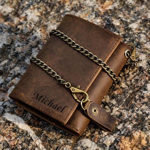 May include: A brown leather wallet with a chain, featuring the name "Michael" engraved on the front. The wallet has a clasp and a leather strap with a snap closure. The chain is a bronze color.
