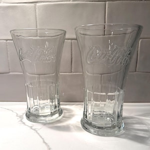 May include: Two clear Coca-Cola glasses. Each glass has a flared shape with a wide base and a slightly narrower top. The Coca-Cola logo is embossed on the front of each glass. The glasses are made of clear glass and are sitting on a white surface.