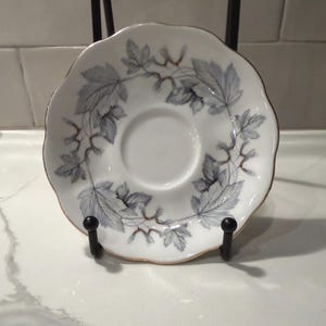 May include: White saucer with a gray leaf and vine pattern encircling the center. The rim has a gold trim. The saucer is displayed on a black metal stand.