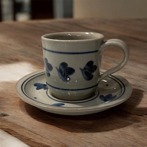 May include: A ceramic teacup and saucer set. The cup and saucer are light gray with blue floral designs and blue stripes. The cup has a handle. The set is on a wooden surface.