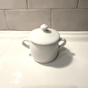 May include: A small, white ceramic sugar bowl with a matching lid. The bowl has two small handles and a rounded shape. The lid has a small knob on top. The sugar bowl is sitting on a white surface.