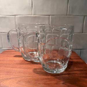 Könnte beinhalten: Zwei Bierkrüge aus klarem Glas mit einer traditionellen, genoppten Außenstruktur und einem stabilen Henkel. Die Krüge sind transparent, haben einen dicken Boden und einen abgerundeten Körper für den Ausschank von Getränken.