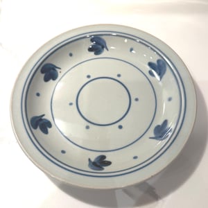 May include: A round, off-white ceramic plate with a blue floral design. The plate features a blue border, a central circle, and stylized blue leaf motifs. Dots are evenly spaced around the center circle and the outer edge.