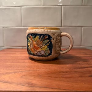 May include: A ceramic mug with a beige base and a prominent relief design featuring a violin and musical instruments in shades of yellow, green, and blue against a dark blue background. The mug has a textured handle and a decorative border.