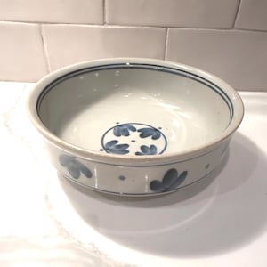 May include: A ceramic bowl with a white interior and exterior. The rim is accented with two thin blue lines. The exterior features blue floral designs. The bowl is round and sits on a white surface.