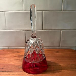 May include: A clear and deep red glass bell featuring a diamond-cut pattern on the upper section and a smooth, solid red base. The handle is made of clear glass, extending upward from the top of the bell, which has a flared, circular rim.