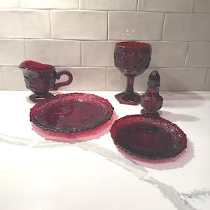 Könnte beinhalten: Eine Sammlung rubinroter Glaswaren, darunter ein Sahnekännchen, ein Weinglas, zwei Teller und ein kleiner Behälter mit Deckel. Die Teile haben ein detailliertes, geprägtes Muster und sind auf einer weißen Oberfläche angeordnet.
