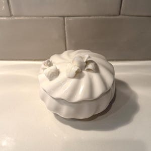 May include: A white ceramic trinket box with a swirled design and small floral accents on the lid. The box has a glossy finish and a round shape, suitable for storing small jewelry items or keepsakes.