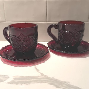 Könnte beinhalten: Zwei rubinrote Teetassen und Untertassen aus Glas. Die Tassen und Untertassen haben ein strukturiertes, verziertes Design. Die Tassen haben Henkel und stehen auf passenden Untertassen. Das Set wird auf einer weißen Oberfläche präsentiert.
