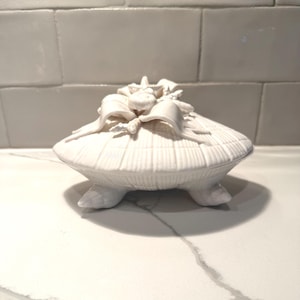以下が含まれることがあります： 白いセラミック製の貝殻型小物入れ。蓋にはリボンと花の装飾が施されています。箱は3つの小さな足の上にあります。表面はリブ加工が施されています。