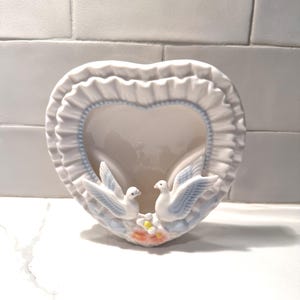 Può includere: Una tasca da parete a forma di cuore in ceramica bianca con bordo a balze e rifiniture azzurre. Due colombe bianche con ali azzurre sono appollaiate su una base floreale. Un pezzo decorativo per l'arredamento della casa.