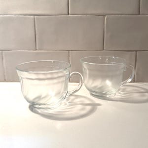 May include: Two clear glass teacups with handles. The cups have a rounded shape with a slight swirl design on the lower half. They are sitting on a white surface in front of a white tiled wall.
