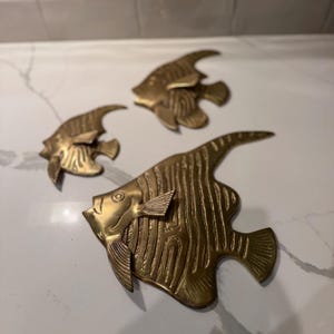 May include: A set of three brass fish wall decorations in varying sizes. Each fish features a polished metallic finish with engraved vertical line patterns on the body, distinct fins, and a tail, creating a cohesive set of nautical wall art pieces.