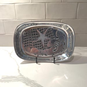 May include: An oval silver-toned metal tray featuring a raised relief design of a starfish, various seashells, and a fishing net pattern. The tray has a polished, reflective surface and is displayed upright on a small black wire stand.