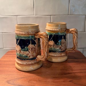 May include: Two ceramic beer steins featuring a relief design of a village scene with houses and trees. The mugs have a cream-colored base, a dark blue background, and textured handles that resemble twisted wood, with decorative borders at the top and base.