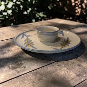 Op de afbeelding: Een keramische theekop en schotel set. De gebroken witte set heeft blauwe en gele bladeren. De kop staat in het midden van de schotel. De rand van de kop en de rand van de schotel zijn blauw. De set staat op een houten oppervlak.