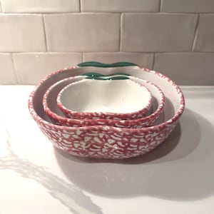 May include: Set of three ceramic bowls nested together. The bowls are white inside with a red and white speckled pattern on the outside. Each bowl has a green leaf-shaped handle. The bowls are designed to look like apples.