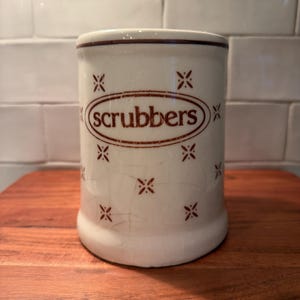 May include: A cylindrical ceramic container with a cream-colored crackle glaze finish. The exterior features a brown oval border containing the text "scrubbers" in lowercase letters, surrounded by a repeating pattern of small brown star-like motifs.