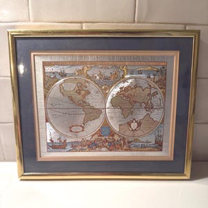 May include: Framed vintage world map print with a gold-coloured frame and a dark blue mount. The map features detailed illustrations of continents, oceans, and sailing ships. The text "NOVA TOTIUS TERRARUM ORBIS GEOGRAPHICA AC HYDROGRAPHICA TABULA" is visible.