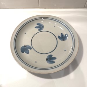 May include: A ceramic saucer with a cream-colored base and a blue floral design. The saucer has a blue border and a central circle, with blue flower motifs and small dots. The design is simple and elegant.
