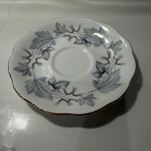 May include: White saucer with a scalloped edge and a gold rim. The saucer features a delicate pattern of gray leaves and vines around the edge. The center is plain white, creating a classic and elegant design.